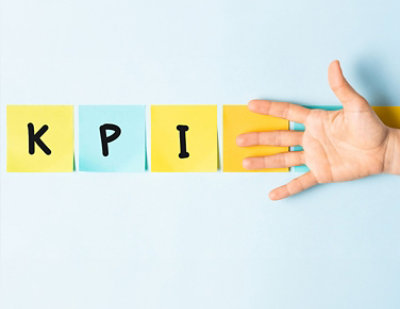 Four sticky notes spell out "KPI" with a hand placed next to them on a light blue background.