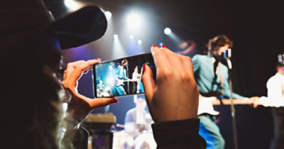 Osoba horizontalno drži pametni telefon i snima nastup uživo muzičke grupe na sceni, a u pozadini se vide scenska svetla.