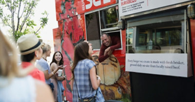 Muškarac se naginje kroz prozor kamiona koji prevozi hranu da bi uručio obrok nasmejanom klijentu, dok drugi ljudi čekaju u redu; tekst na kamionu