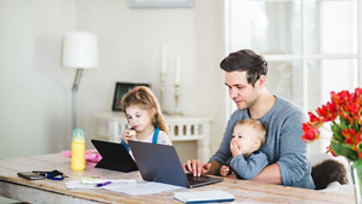 Mężczyzna pracuje na laptopie przy stole z niemowlęciem na kolanach i małym dzieckiem obok niego korzystającym z innego laptopa. Na stole leżą również klucze, butelka i dokumenty.