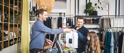 Muškarac za pultom u maloprodajnom objektu dodaje mladoj devojci korpu za kupovinu. Pored nje stoji drugi muškarac, verovatno čuvar.