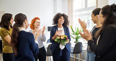 Žena odevena u odelo drži buket cveća i osmehuje se, a grupa ljudi oko nje aplaudira i kliče