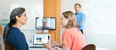 Due donne sedute alla scrivania con un portatile, impegnate in una conversazione. Un uomo in piedi sullo sfondo accanto a una lavagna elettronica con note