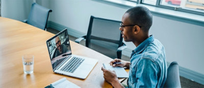 Una persona seduta a un tavolo di legno, impegnata in una videochiamata sul portatile. Ha un blocco note e una penna in mano