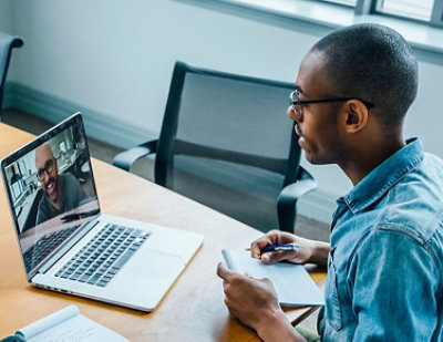 A person in a denim shirt sits at a conference table with a notebook, having a video call on a laptop.