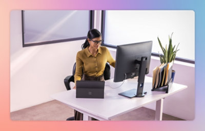 A computer on a desk in front of a window with a person wearing a yellow shirt.