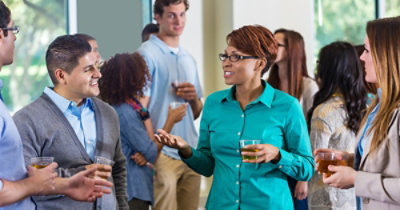 Grupa formalno odevenih osoba koje međusobno razgovaraju i ispijaju piće na društvenom događaju u zatvorenom prostoru.