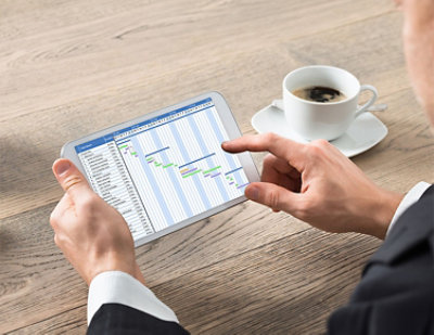 A person pointing at a tablet displaying a Gantt chart with a cup of coffee on the wooden table nearby.