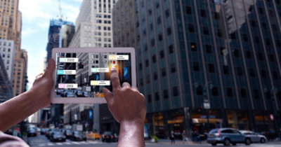 Osoba drži tablet na čijem ekranu se vidi pejzaž grada i prikazuju se informacije proširene stvarnosti o različitim zgradama
