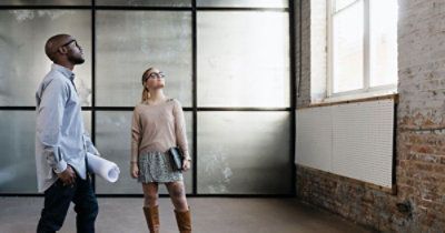 Dve osobe stoje u prostoriji opremljenoj u industrijskom stilu i gledaju ka prozoru. Jedna od njih drži nacrte planova