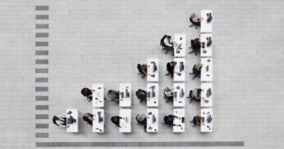 Pogled odozgo na kancelarijske službenike koji rade za izduženim stolovima poređanim u pravilnim redovima na krupno popločanom podu.