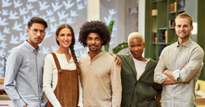 Pet osoba stoje jedna pored druge i smeju se u jarko osvetljenoj prostoriji sa ukrasnim šarama na zidu