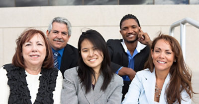 Raznovrsna grupa petoro ljudi u poslovnoj odeći, sede ispred zgrade i smeju se.