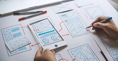 Osoba crta na papiru modele veb sajta i rasporede aplikacija, sa različitim skicama i strelicama koje pokazuju tok