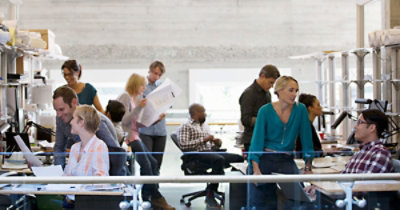 Užurbana scena u kancelariji u kojoj devet osoba rade za stolovima, sarađuju na projektima i pregledaju dokumente