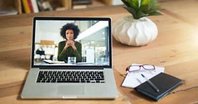 Un portatile su una scrivania in legno visualizza una riunione virtuale con una donna. Accanto, ci sono occhiali, un quaderno e una penna