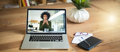 A laptop on a wooden desk displays a virtual meeting with a woman. Nearby, there are glasses, a notebook with a pen