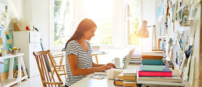 Osoba pracuje na laptopie przy biurku zagraconym książkami i papierami w jasnym, nasłonecznionym pomieszczeniu.