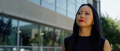 Person with long dark hair in a sleeveless top standing outside a modern glass building with greenery.