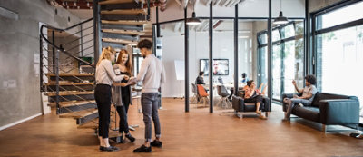 People are standing and talking near a staircase in a modern, open office space with large windows.
