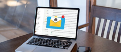 Laptop na drewnianym stole wyświetla otwartą skrzynkę odbiorczą z nałożoną ikoną koperty i plakietką powiadomienia.