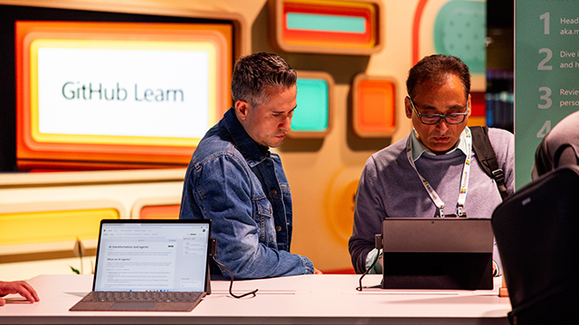 Two people at a “GitHub Learn” booth use laptops at a demo counter, with colorful display panels in the background.