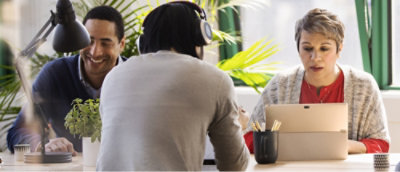 Two individuals working on laptops at a desk with plants in the background.