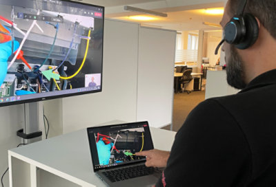 A man wearing headphones works on a laptop at a standing desk in an office, displaying robotics programming on his screen.