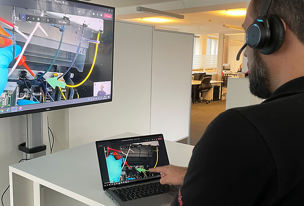 Een man die een hoofdtelefoon draagt en aan een staand bureau op een laptop werkt in een kantoor met robotica-programmering op zijn scherm.