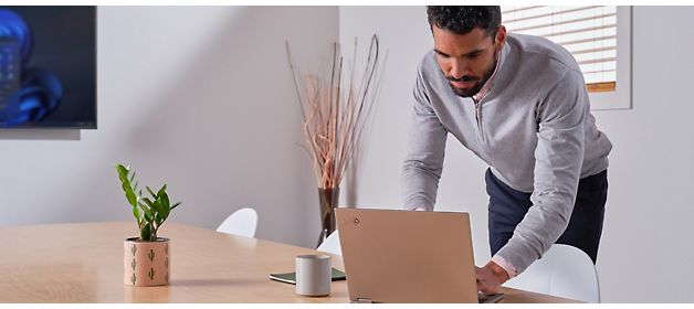 Mężczyzna w szarym swetrze stoi i pochyla się nad laptopem leżącym na drewnianym biurku w jasnym biurze.