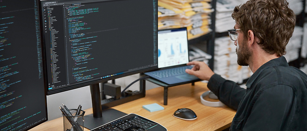 一名男子在办公室环境中使用双显示器显示代码和图形，在计算机上分析数据。