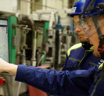 Two factory workers wearing safety gear are discussing information displayed on a screen in an industrial setting.