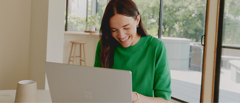 whiteboard presentation online A person in a green sweater smiles while using a laptop at a table in a bright room with large windows.