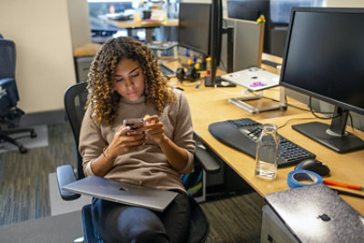 Una persona con i capelli ricci è seduta alla scrivania dell'ufficio intenta a guardare il suo smartphone. Più monitor di computer e articoli da ufficio sono visibili sulla scrivania.