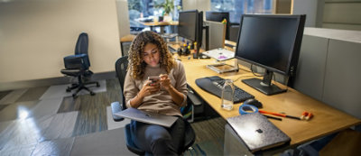 A person with curly hair is seated at an office desk, looking at a smartphone. Multiple computer monitors and office supplies are visible on the desk.