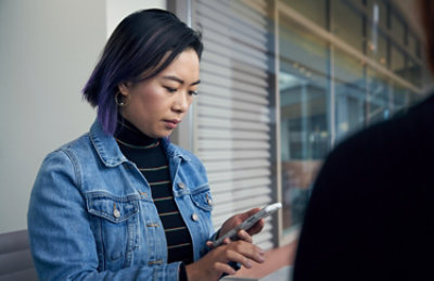 Persona con capelli corti indossa una giacca jeans guarda in basso al telefono in un ambiente interno con persiane sullo sfondo.