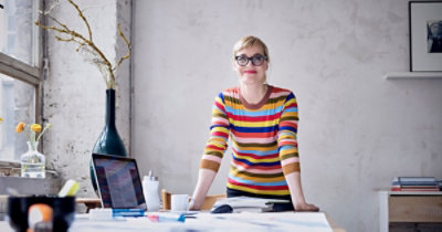Žena sa naočarima odevena u prugasti džemper stoji u dobro osvetljenoj prostoriji za stolom na kojem se nalaze razbacani papiri, laptop i vaza.