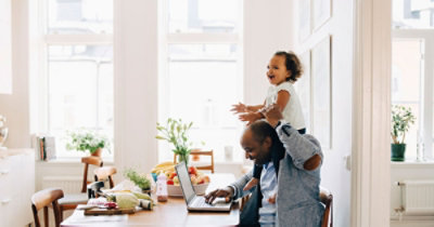 A person holding a baby on his shoulders while sitting at a table with a computer