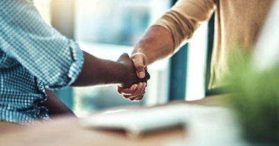 Dve osobe se rukuju u zatvorenom prostoru, jedna od njih ima na sebi kariranu košulju dugih rukava, a druga džemper bež boje