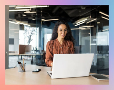 A person with curly hair, wearing glasses and a brown polka dot shirt, sits at a desk using a laptop in a modern office setting.