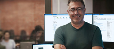 Un hombre delante de dos escritorios de pantalla grande y sonriendo.