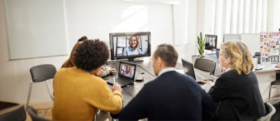 Kilka osób siedzi przy stole konferencyjnym, prowadząc rozmowę wideo, z jedną osobą widoczną na dużym ekranie i drugą na laptopie. W tle widać roślinę.