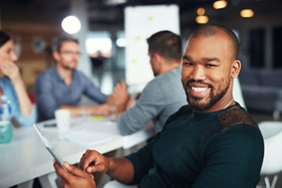 Un uomo che sorride in primo piano con tre persone sfocate sullo sfondo sedute intorno a un tavolo in una stanza luminosa.