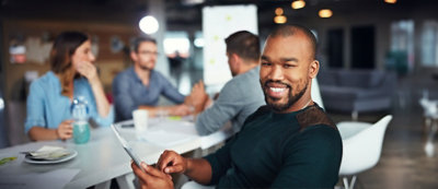 A smiling man in the foreground with three people blurred in the background sitting around a table in a brightly lit room.