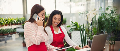 Dve žene rade zajedno u rasadniku. Jedna razgovara telefonom i drži fasciklu, a druga koristi laptop