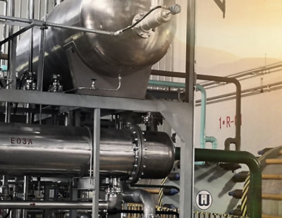 A large silver cylinder with pipes in a factory, labeled R-E03A H, with a person on a green railing nearby.