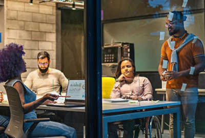 Team collaborating in a modern office, discussing charts at a table with laptops, notes on glass walls, and shared workspace setting.
