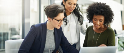Three women working on one laptop in office discussing something