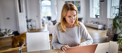 Una donna con capelli lunghi è seduta al tavolo di una stanza luminosa e moderna, concentrata sul portatile davanti a lei.