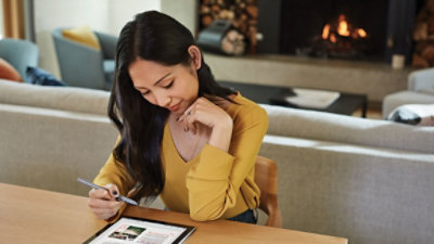 Žena u žutoj majici bez rukava sedi za stolom i koristi olovku na tabletu, a u pozadini se nalazi kamin i sofa.